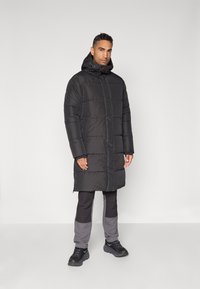 Doudoune noire avec capuche, présentant des coutures horizontales, une fermeture éclair à l'avant et des poches latérales. Portée sur un pantalon noir et gris et des baskets.