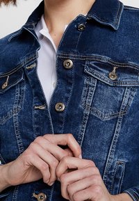 Modrá džínová bunda tmavě modré barvy se stříbrnými knoflíky, se čtyřmi kapsami a klasickým límcem. Má strukturovanou látku a střih na míru.