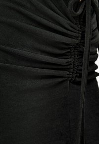 Prenda de tejido negro con textura recogida, que presenta un sutil acanalado y un detalle de nudo lateral. Material suave y elástico con un acabado liso.