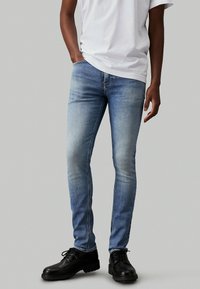 Jeans de mezclilla azul ajustados con un acabado desgastado, detalles de costura y un corte de tiro medio, llevados con una camiseta blanca lisa y zapatos negros.