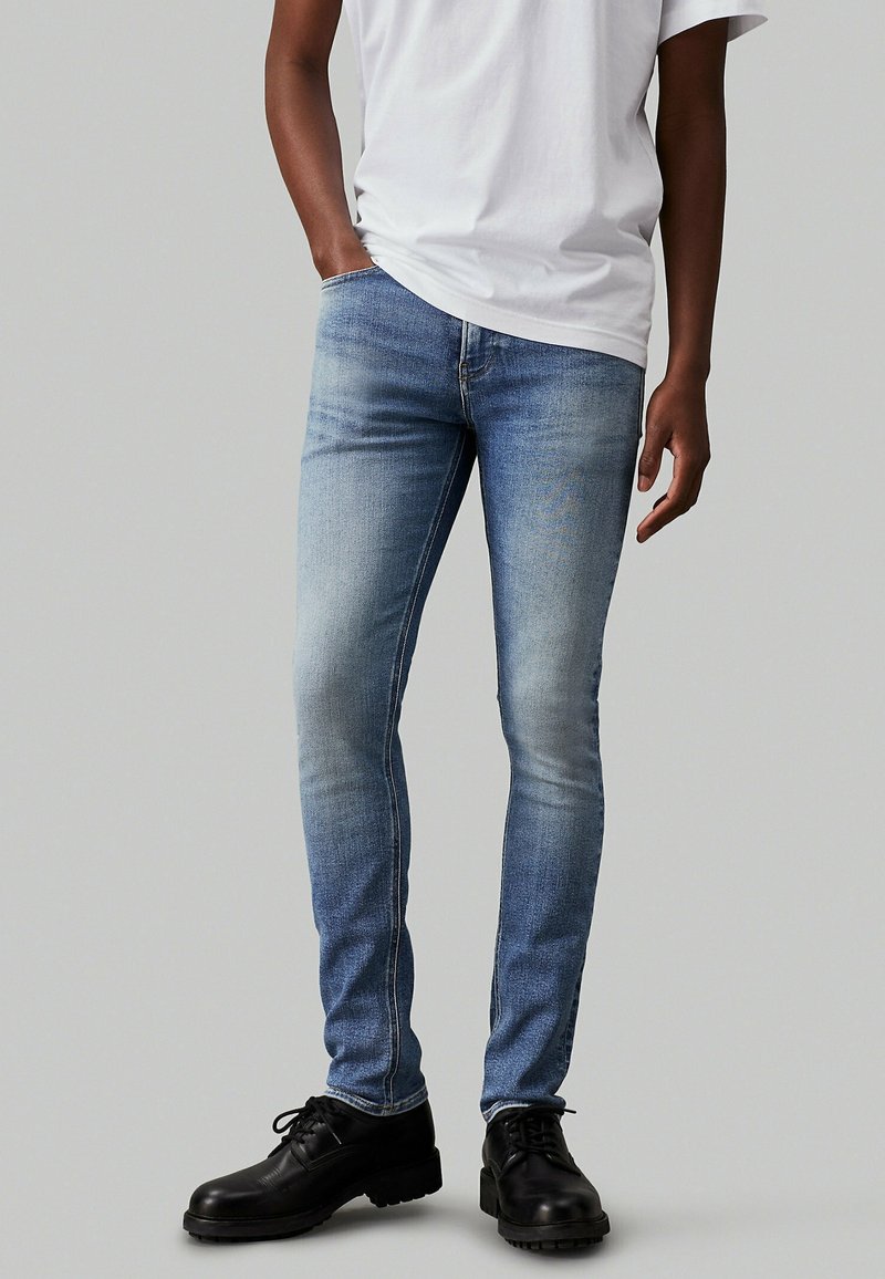 Jeans de mezclilla azul ajustados con un acabado desgastado, detalles de costura y un corte de tiro medio, llevados con una camiseta blanca lisa y zapatos negros.