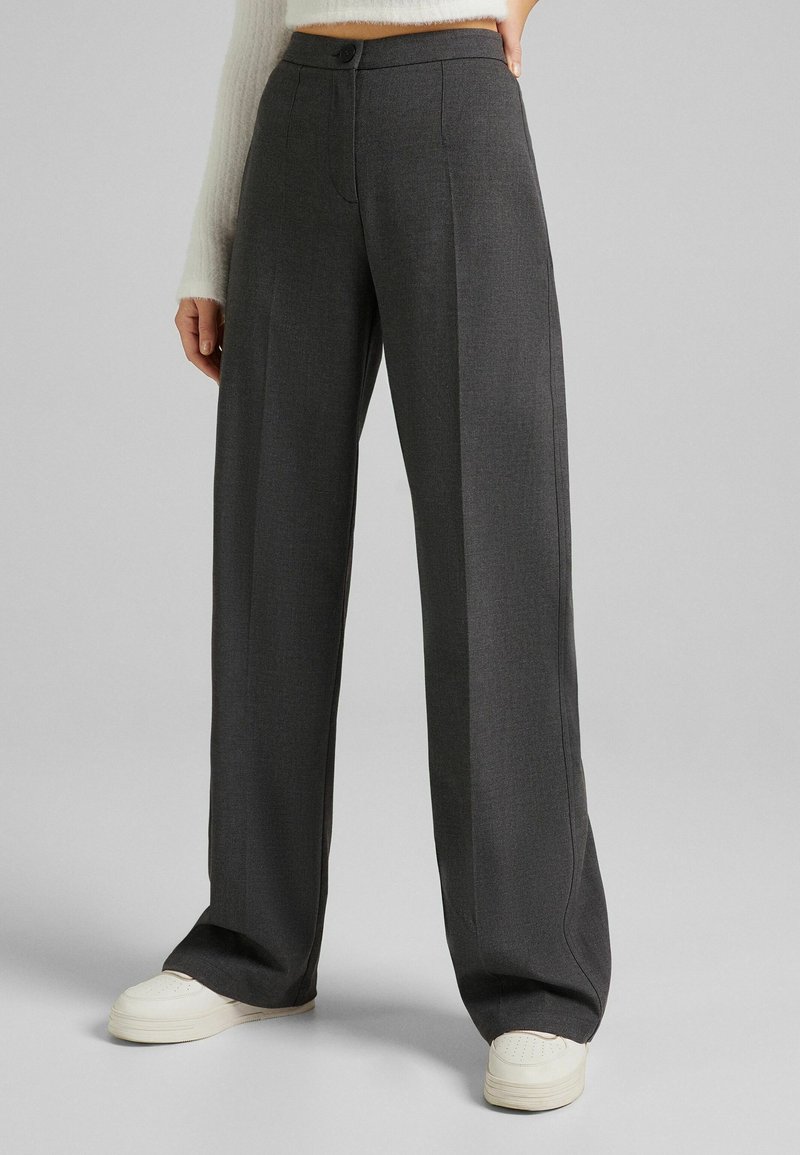 Pantalon large gris foncé porté avec des baskets blanches et un pull blanc court et duveteux sur un fond gris uni.