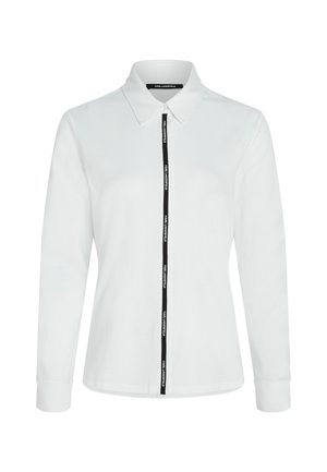 Balta garām piedurknēm kreklā ar apkakli, ar melnu rāvējslēdzēju akcentu un logotipa lentes dizainu priekšpusē. Izgatavots no mīksta kokvilnas materiāla.