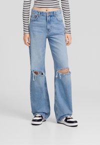 Jeans in denim azzurro chiaro con vita alta, gambe larghe e grandi strappi sulle ginocchia. Il tessuto sembra morbido con una texture leggermente sbiadita.
