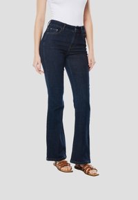 Flared denim jeans in donkeblauwe kleur, met een hoge taille en stikselaccenten. Gecombineerd met bruine sandalen met bandjes en een wit topje.