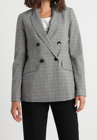 Blazer à carreaux gris croisé avec deux grands boutons noirs, revers crantés et poches latérales. Le modèle porte une chemise noire et un pantalon.