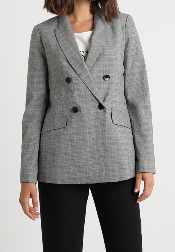 Blazer à carreaux gris croisé avec deux grands boutons noirs, revers crantés et poches latérales. Le modèle porte une chemise noire et un pantalon.