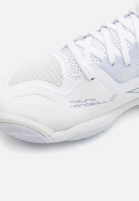 Pantof de handbal Mizuno în alb, cu secțiuni din plasă, suprafață texturată, șireturi plate și detaliu "Dura Shield" pe talpă pentru durabilitate și aderență.