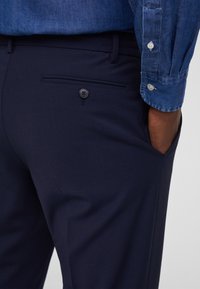 Pantalones entallados de color azul marino, confeccionados en una tela suave, con un bolsillo trasero con detalle de botón y un bolsillo lateral, combinados con una camisa de mezclilla.
