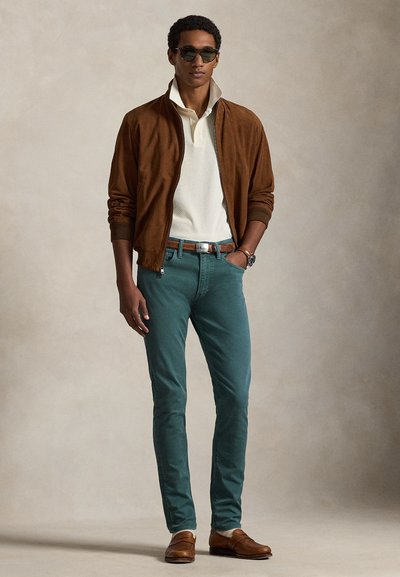 Brun semsket bomberjakke, kremfarget strikkepolo, turkise slanke bukser, brun skinnbelte, brune loafers og mørke solbriller.