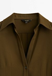 Chemise marron à col avec un seul bouton visible et une étiquette de marque à l'intérieur de l'encolure.