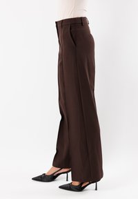 Pantaloni larghi marroni con pieghe frontali molto definite, una texture liscia e un design formale, abbinati a decolletè nere a punta.