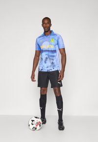 Futbalový dres Puma obsahuje svetlomodrý dres s grafickým dizajnom, čierne šortky, čierne ponožky a futbalovú loptu s čierno-bielym vzorom.