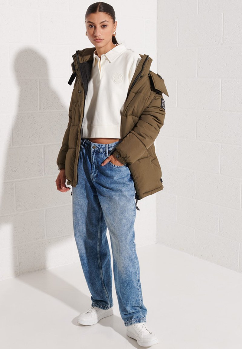 Chaqueta de plume de color marrón, sudadera blanca corta y jeans sueltos de color azul claro. Lleva zapatillas blancas y está estilizada con accesorios mínimos.