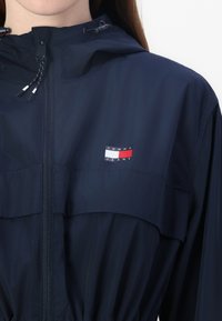 Giacca a vento blu navy con cappuccio regolabile, zip frontale e vita elasticizzata. Presenta un piccolo logo Tommy Jeans in rosso, bianco e blu.