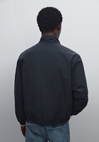 Black windbreaker jacket with a high collar, elastic cuffs, and hem. Smooth, lightweight fabric; back view shows minimalist design.