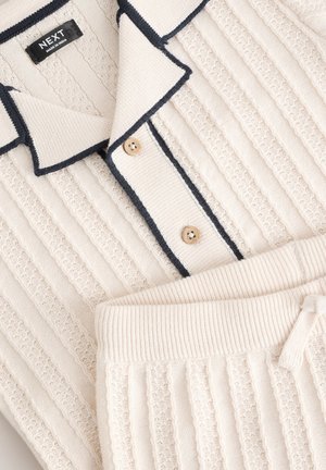 Kerrosleikattu neulosmukavuusasu kermansävyinen, jossa on kauluksellinen yläosa laivastonsinisin somistein ja kaksi nappia, yhdistetty samanlaisiin elastiseen vyötäröön sopiviin housuihin.