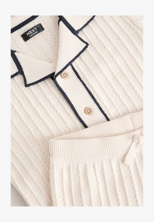 Kreminės spalvos ribbinta megzta namų drabužių komplektas, kurį sudaro apykaklėmis puoštas viršus su tamsiai mėlyna apdaila ir dviem mygtukais, kartu su atitinkančiais elastinės juostos kelnais.