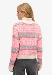 Pull en tricot avec un col haut, motifs en losanges rose et gris, et poignets côtelés. Le matériau semble doux et texturé.