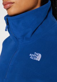 Niebieska polarowa kurtka z wysokim kołnierzem i półzamkiem. Posiada białe, haftowane logo "The North Face" na piersi. Miękka tekstura.