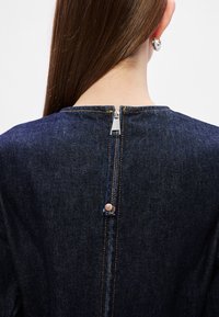 Σκούρο τζιν φόρεμα με ψηλό λαιμόκοψη, που διαθέτει ορατό μεταλλικό φερμουάρ και ξύλινο κουμπί στην πλάτη, μινιμαλιστική ραφή.