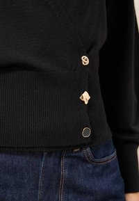 Pulover negru din lână tricotat cu margine ribbed, cu trei butoane decorative aurii, purtat cu blugi denim albastri cu ocheade portocalii.
