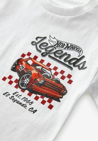 Tricou alb cu un grafic roșu al unei mașini Hot Wheels, accentuat cu motivos cu carouri, textul "Legends" și "Est. 1968 El Segundo, CA" imprimat dedesubt.