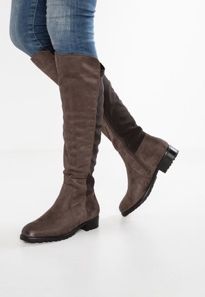 Bottes en daim marron mi-mollet portées avec un jean bleu, un pied levé montrant la fermeture éclair latérale de la botte et son talon bas.