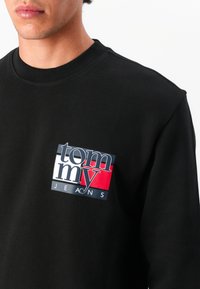 Zwarte sweatshirt met een geribbelde halslijn en een rechthoekig logopatch in blauw, wit en rood, met de tekst "tommy JEANS."