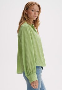 Hellgrünes Hemd mit Knopfleiste und strukturiertem, gefälteltem Design. Verfügt über gekrempelte lange Ärmel und einen spitzen Kragen. Getragen mit blauen Jeans.