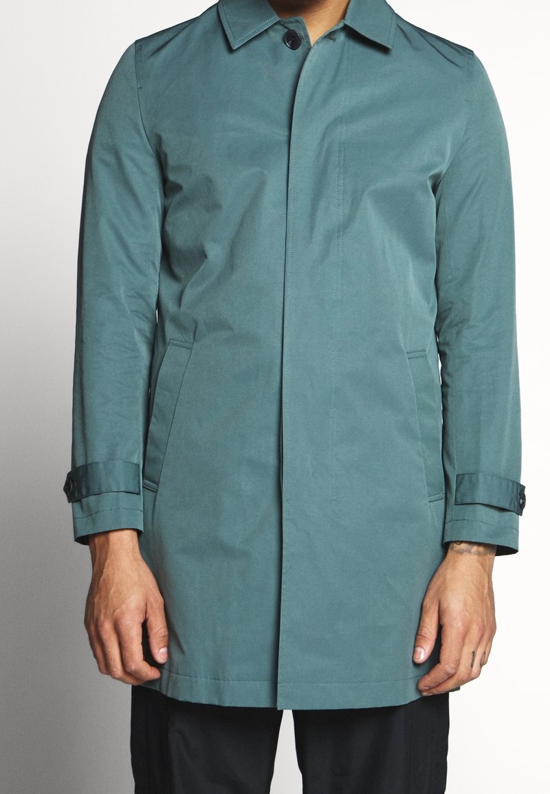 Homme portant un long manteau bleu sarcelle avec des brides boutonnées aux poignets et des poches latérales, debout sur un fond uni.