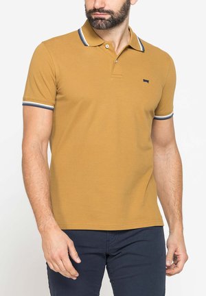 Uomo che indossa una polo giallo senape a maniche corte con colletto e bordi delle maniche a righe blu navy e bianche, abbinata a pantaloni blu navy scuro.