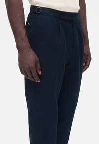 Pantalon bleu foncé sur mesure avec boucle latérale réglable et fermeture par bouton, porté par une personne à la peau foncée et au top clair.