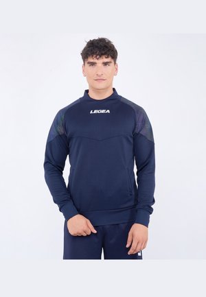 Giovane uomo con capelli ricci indossa una felpa blu navy a maniche lunghe con spalle decorate e logo "LEGEA" sul petto, in piedi su sfondo bianco.