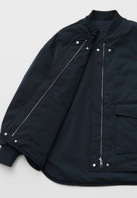 Schwarze Steppjacke mit silbernem Reißverschluss und Druckknöpfen, offen, um das Innenfutter zu zeigen, mit gerippten Bündchen und einer vorderen Tasche mit Klappe.