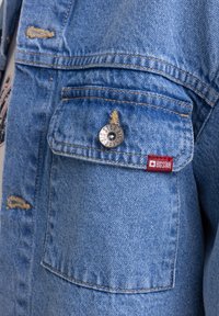 Giacca in denim azzurro chiaro con dettagli consumati, un bottone a scatto argentato e un'etichetta in tessuto rosso con il nome del marchio sulla tasca.