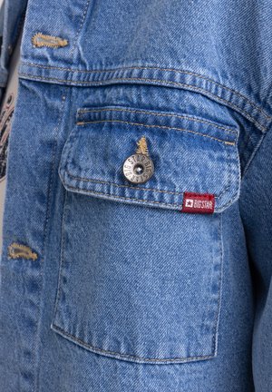 Svetlo modra jeans jakna z obrabljenimi detajli, srebrnim zapiranjem na gumb in rdečo tkaninsko oznako z imenom blagovne znamke na žepu.