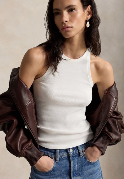 Γυναίκα που φοράει λευκό ριμπ χωρίς μανίκια τοπ, μπλε τζιν και καφέ δερμάτινο μπουφάν περασμένο στους ώμους, με σκουλαρίκια κοχύλι, χέρια στις τσέπες.