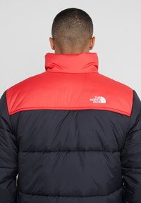 Giacca a vento con sezione superiore rossa e sezione inferiore blu navy, caratterizzata da una texture trapuntata orizzontale e un logo bianco sul retro.