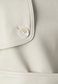 Crème-kleurige neplederen jas met een grote ronde knoop, prominente stiksels en een gestructureerde kraagdetail.