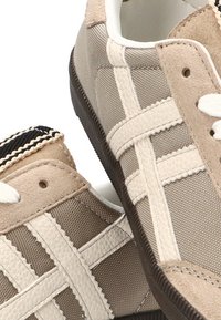Zapatillas beige hechas de cuero y tejido texturizado, con un patrón de cruz, cordones blancos y una suela de goma con una franja en contraste.
