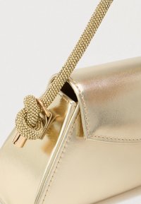 Arany fémes kézitáska, sima, szögletes formával. Textúrált pánttal rendelkezik, amelyen csomó részlet található, és fényes kiegészítők díszítik.