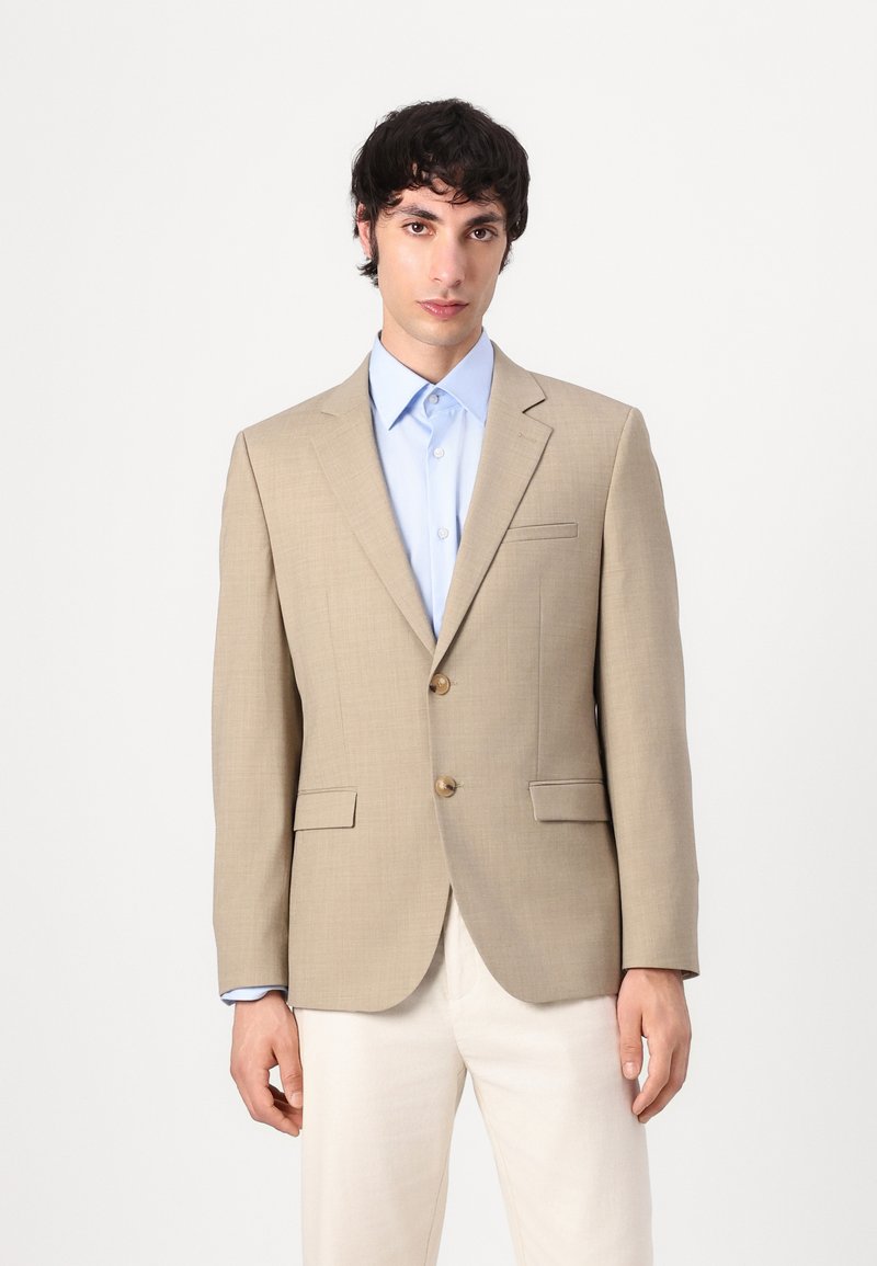 Beige getailleerde blazer met twee gouden knopen, notch revers en twee voorzakken, gedragen over een lichtblauw shirt, gecombineerd met lichtgekleurde broeken.
