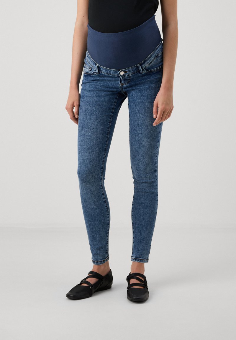 Jeans ajustados de denim azul oscuro con una cinturilla alta y elástica de color marino. Llevados con zapatos negros planos con tiras cruzadas.