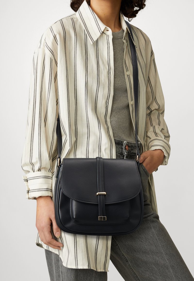 Persona che indossa una camicia a righe color crema, un top grigio e jeans, con una borsa a tracolla in pelle nera con patta frontale e dettaglio in oro.