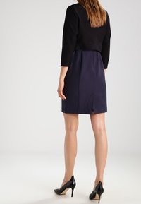 Svart kort blazer ovanpå en marinblå klänning med en liten slits bak. Glansiga svarta högklackade skor fulländar outfiten. Mjuk tygstruktur.