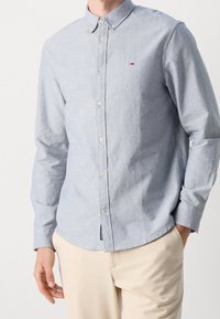 Muž oblečený v bleděmodré košili s dlouhým rukávem a malým logem na hrudi a béžových kalhotách, s jednou rukou v kapse, stojící na neutrálním pozadí.