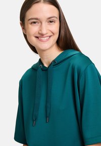 Turkusowa bawełniana bluza z kapturem na sznurku, krótkimi rękawami i gładką fakturą, prezentująca luźny krój i minimalne wykończenie.