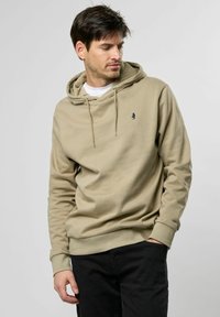 Beige hoodie gemaakt van zachte stof, met een trekkoordkap en een klein logo op de borst. Gepaard met zwarte broeken.