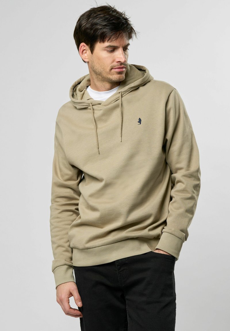 Beige hoodie gemaakt van zachte stof, met een trekkoordkap en een klein logo op de borst. Gepaard met zwarte broeken.